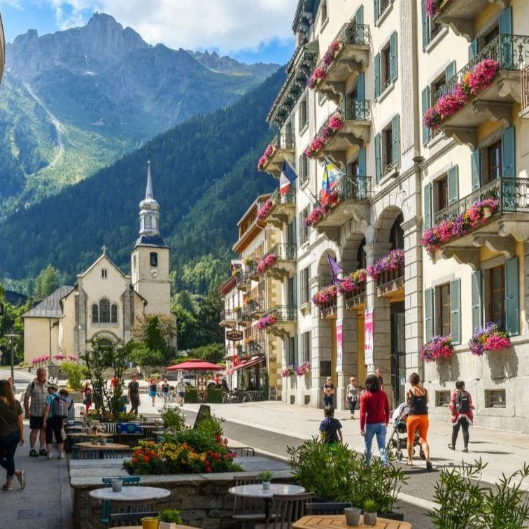 Place du Triangle de l'Amitié - Chamonix Mont-Blanc Rue animée avec des bâtiments fleuris, une église et des montagnes en arrière-plan.
