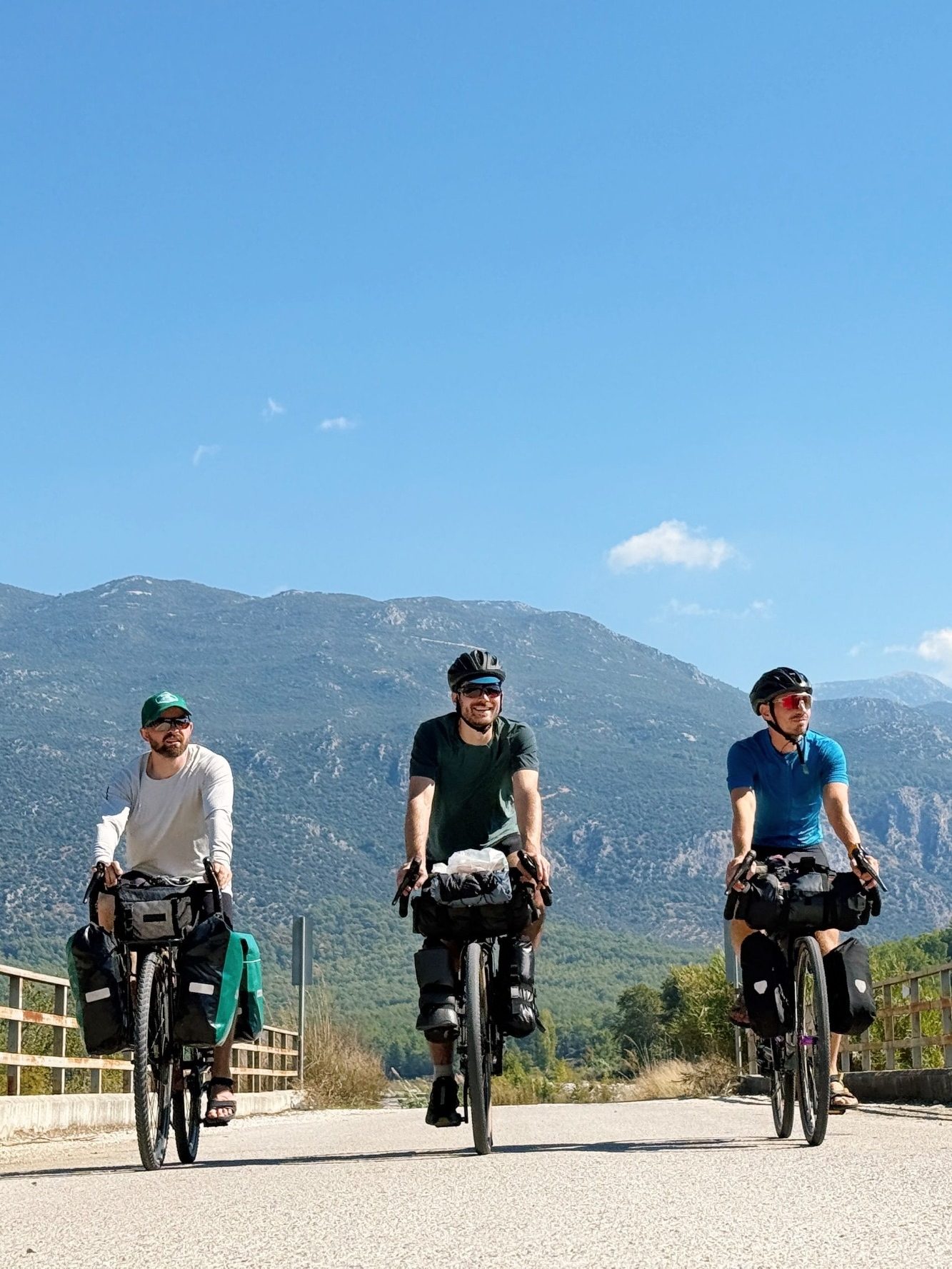 Portage Bagages randonnée à vélo avec VTC-Haute-Savoie Trois cyclistes avec des sacs de vélo sur une route entourée de montagnes.