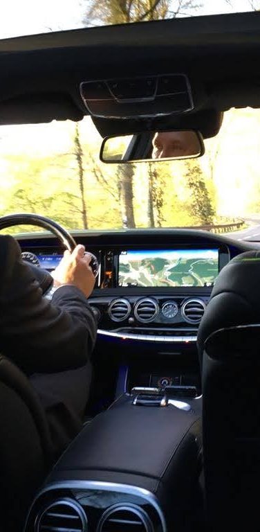 Taxi VTC Haute-Savoie by Classe-Affaires VTC - Chauffeur privé de sécurité - France - Suisse - Luxembourg Homme en costume conduisant une voiture avec un ciel dégagé visible à travers le toit.
